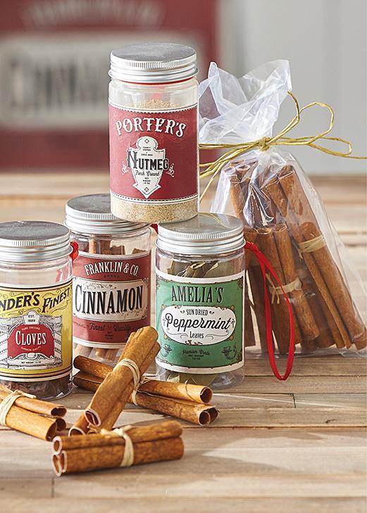 Four spice container ornaments are arranged on a wooden board. Cloves, cinnamon, and peppermint, with Nutmeg stacked on top. Loose and packaged cinnamon sticks are scattered around the display.