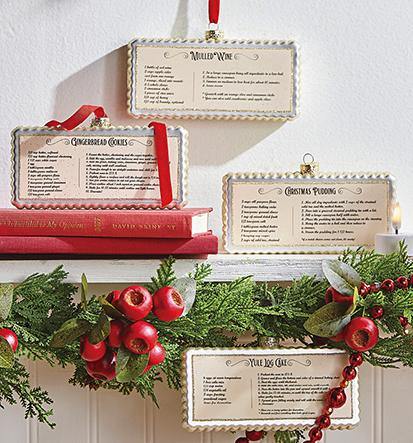 Three rectangular ivory glass ornaments printed with recipes hang above a narrow white mantle decorated with juniper garland , red bead garland,and red berries. A Fourth ornament hangs from the garland. The recipes are Mulled Wine, Gingerbread Cookies, Christmas Pudding, and Yule Log Cake.